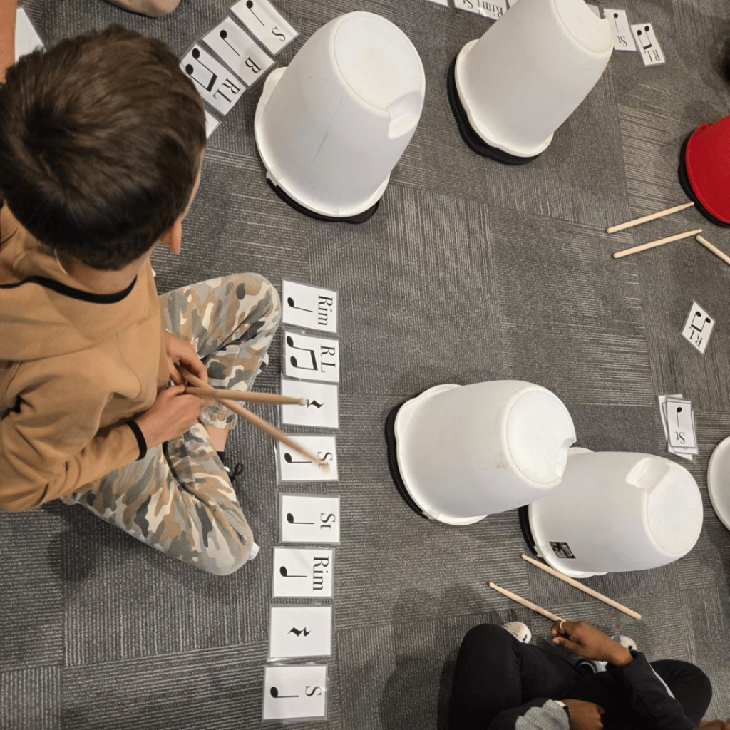 Child participating in a rhythm reading and drumming activity using visual music notation cards and plastic buckets during a Nova Music School percussion class.